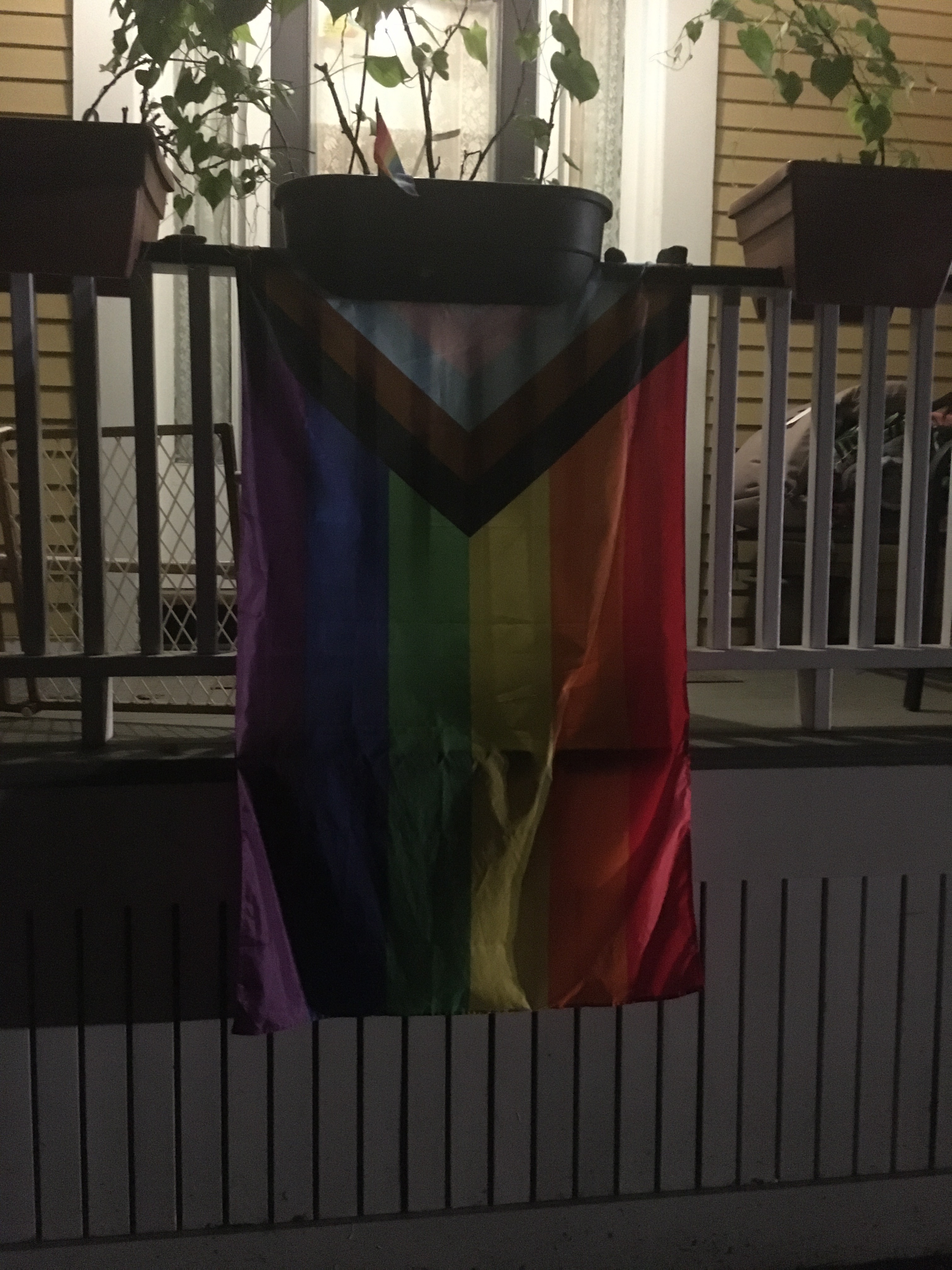 New Trans Flag update to the rainbow gay flag at dusk hanging from a porch